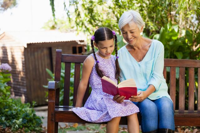 Smiling grandmother reading book to granddaughter while sitting on wooden bench at backyard