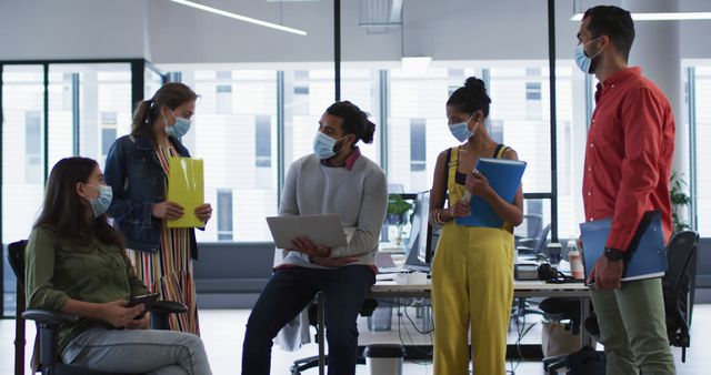Group of young professionals collaborating in modern office while wearing masks and maintaining social distancing. Team members discussing documents and working on laptops. Ideal for showing office teamwork, pandemic precautions in workplaces, and modern work environments.