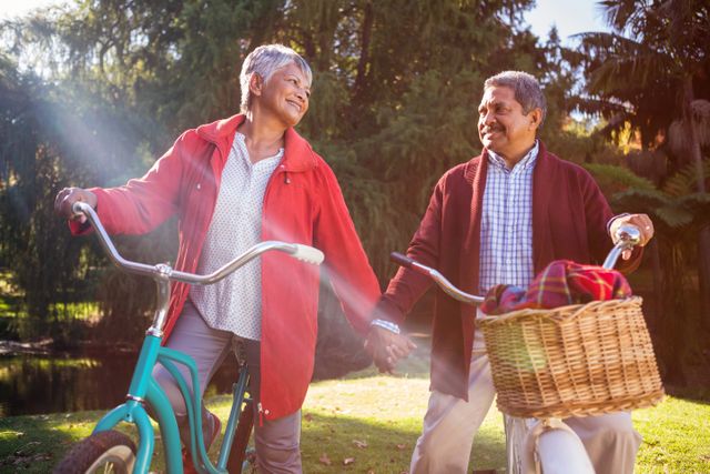 Senior couple enjoying a sunny autumn day in the park with their bicycles. They are holding hands and smiling, showcasing a happy and active lifestyle. This image can be used for promoting healthy living, retirement activities, outdoor recreation, and senior wellness.