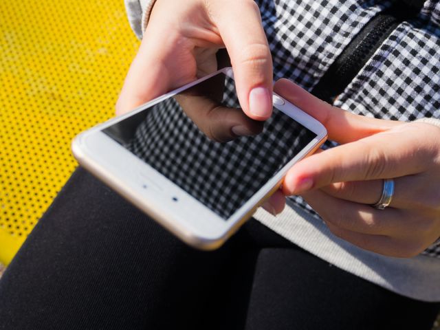Person's hands holding and using a smartphone while sitting on a bright yellow bench. Ideal for websites, blogs, or advertisements related to technology, connectivity, social media, and modern lifestyle.