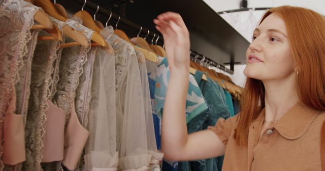 Happy caucasian woman with red hair looking at clothes hanging in shop, copy space. Shopping, fashion, free time, urban living and lifestyle, unaltered.
