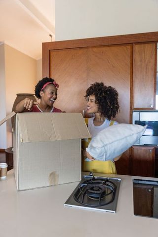 Two friends unpacking moving box in modern kitchen holding pillow and coffee
