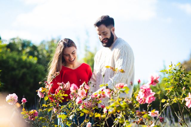 Young couple enjoying a sunny day in the park, smiling and looking at blooming flowers. Ideal for concepts of love, togetherness, happiness, and lifestyle. Perfect for use in advertisements, blogs, social media posts, and articles related to romance, leisure activities, and nature.