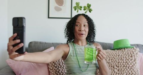 Curly woman taking selfie on couch with shamrock headband and green drink for st patrick's day