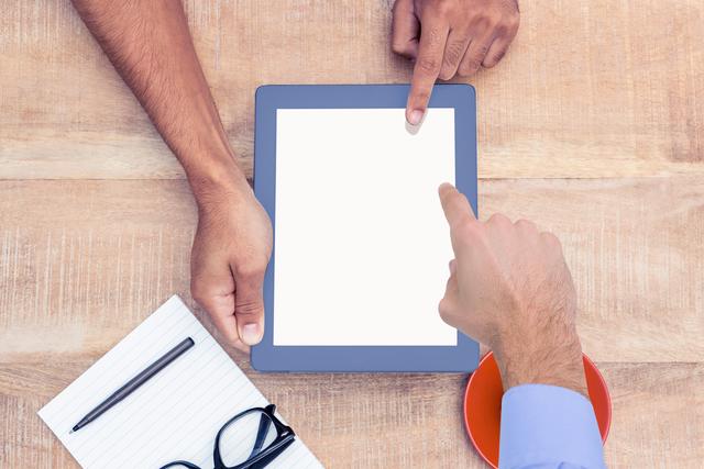 Hands are pointing at a blank tablet screen on a wooden desk, with a notebook, pen, glasses, and a coffee cup nearby. This can be used to illustrate teamwork, meetings, or business planning in a modern office environment. Ideal for websites, blog posts, or advertisements related to office work, technology in business, and collaboration.