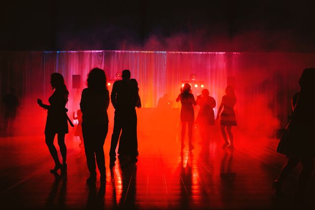 People are dancing on a dark club dance floor illuminated by red lights and surrounded by smoke. This scene captures the energy and atmosphere of a lively nightlife setting, perfect for use in projects related to parties, club events, nightlife promotions, music events, or social gatherings.