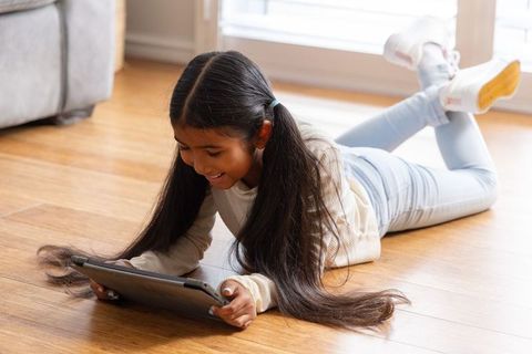 Young girl with long hair lying on hardwood floor using tablet, smiling, casual home learning