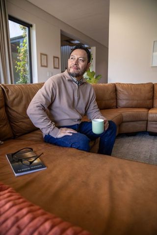 Mature asian man relaxing on leather sofa holding green mug and gazing thoughtfully