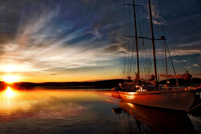 Yacht sits still on calm water reflecting vibrant sunset sky, creating tranquil evening scene. Perfect for travel brochures, vacation advertisements, blogs about sailing or nature, tranquil mood wallpapers, scenic focus visuals.