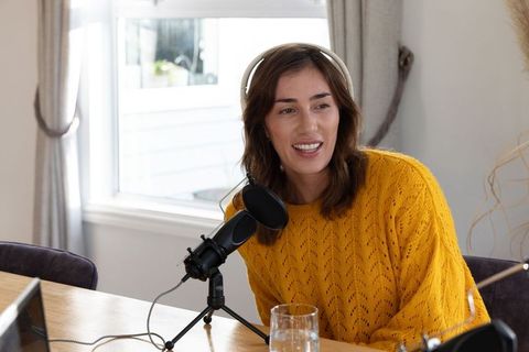 Woman recording cozy home podcast in mustard sweater by window, headphones and microphone