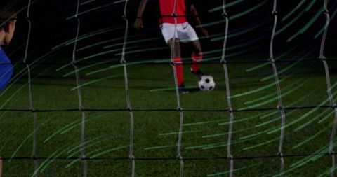Striker preparing penalty kick at night turf through goal net with goalkeeper watching