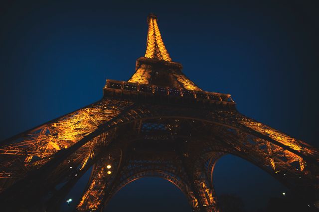 A dramatic night view of the Eiffel Tower with a low angle perspective, highlighting its intricate iron lattice and golden lights against a dark sky. Perfect for use in travel brochures, websites promoting Paris, or editorial content focusing on iconic landmarks.