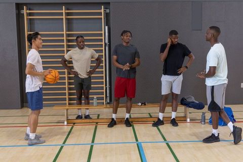 Diverse teammates standing and talking on basketball court holding basketball and bags