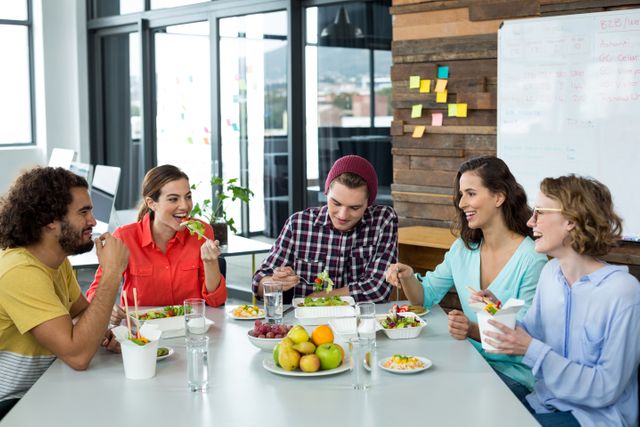 Group of business executives enjoying a meal together in a modern office setting. They are smiling and interacting, suggesting a positive and collaborative work environment. Ideal for use in articles or advertisements about workplace culture, team building, healthy eating at work, or modern office environments.