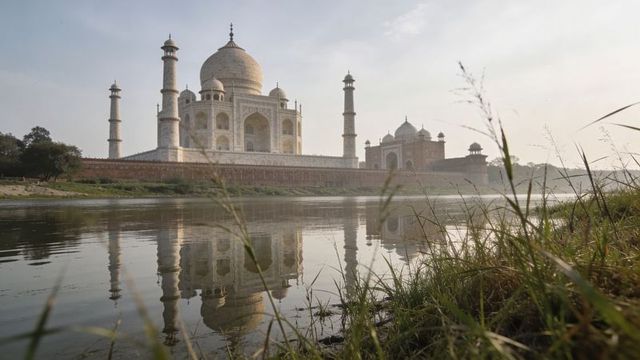 Taj mahal reflecting on river at sunrise with grassy foreground and morning mist