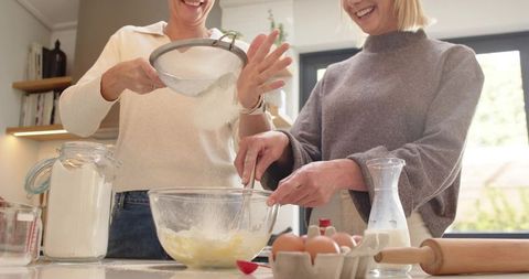 Smiling friends sifting flour and whisking batter at kitchen island for home baking
