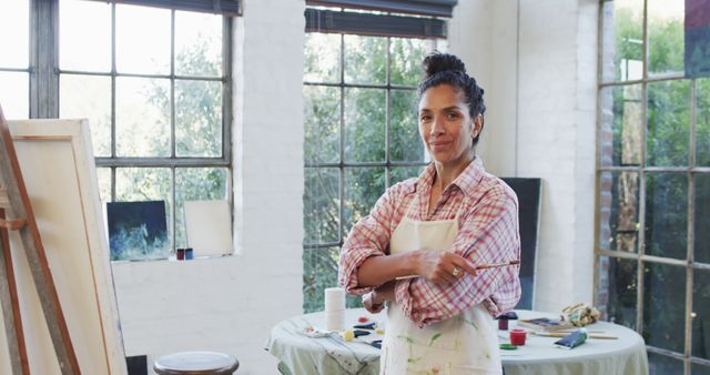 Woman standing in art studio confidently holding paintbrush and wearing apron. Large windows and natural lighting enhance the creative atmosphere. Suitable for use in articles about art, creativity, women in art, personal inspiration, and creative spaces.