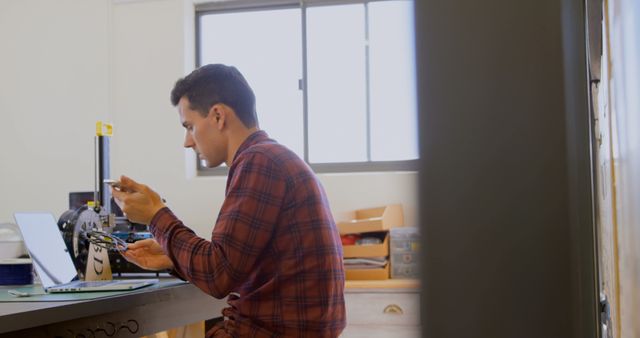 Young man in plaid shirt carefully assembling a 3D printer in a home office setting. He is focused on the task while working on his desk surrounded by tech gadgets and a laptop. Perfect for illustrating concepts of innovation, DIY engineering projects, and modern technology in action. Suitable for use in articles on home tech setups, engineering hobbies, or startup environments.