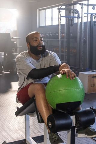 Bearded man resting with green medicine ball on bench in gym, tired athlete recovering