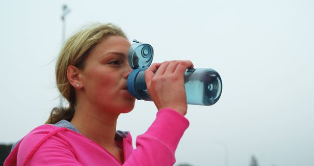 Young woman drinking from blue water bottle while exercising outdoors, showcasing importance of hydration. Useful for health and fitness promotions, hydration campaigns, and active lifestyle advertisements.