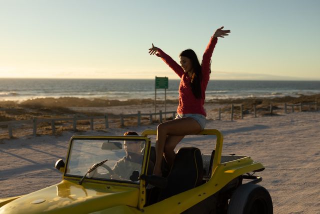 Happy caucasian couple driving beach buggy by sea at sunset, woman sitting on roll bar raising arms. beach stop off on romantic summer holiday road trip.