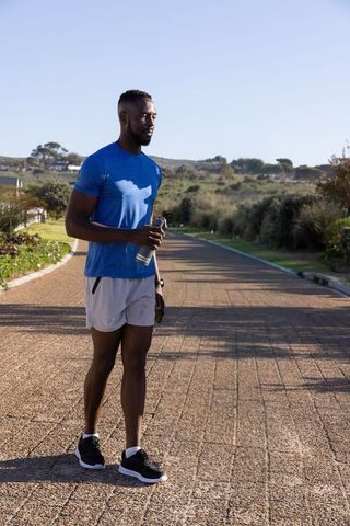 African american man standing on sunlit coastal running path holding water bottle and towel