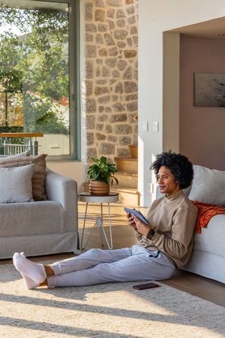 Sunlit living room scene with african american woman sitting on rug holding tablet, phone nearby
