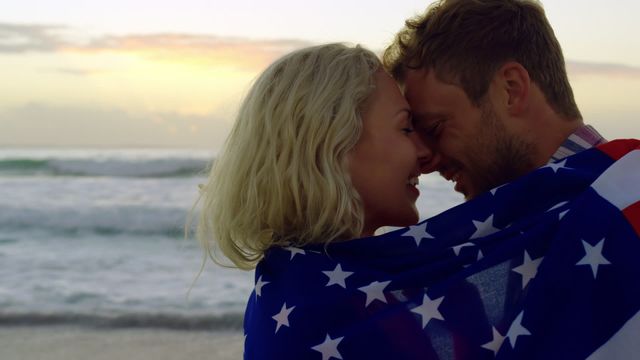 Young Caucasian couple wrapped in American flag, sharing an intimate moment at beach during sunset. They are embracing each other with eyes closed, highlighting themes of love, romance, and patriotism. Perfect for themes related to love stories, national pride, and holiday promotions.