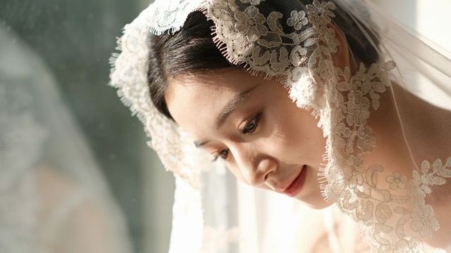 Tilting asian bride wearing embroidered lace veil, lowering eyes in soft window light