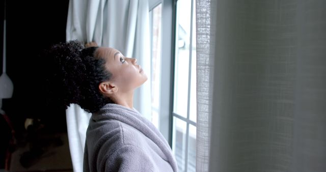 Biracial woman in bathrobe opening curtains in bedroom, copy space. Lifestyle, relaxation and domestic life, unaltered.