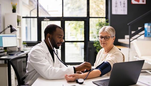 Doctor measuring blood pressure for senior patient at clinic desk with laptop