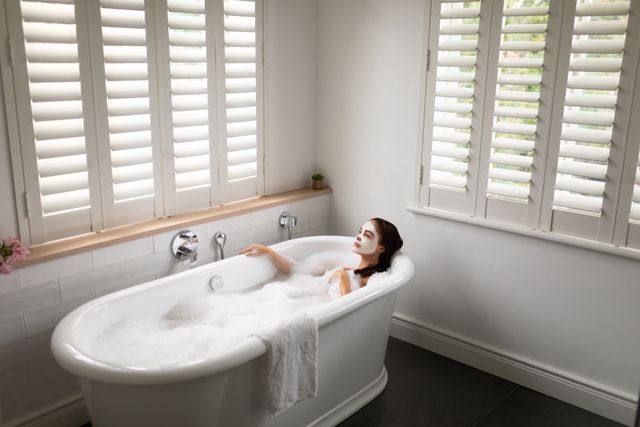 Woman enjoying a bubble bath while wearing a face mask in a modern bathroom with white shutters. Ideal for use in articles or advertisements about self-care, relaxation, home spa treatments, skincare routines, and luxury home interiors.