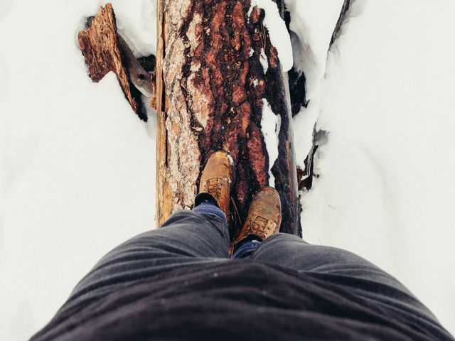 Feet wearing brown boots balancing on fallen tree covered with snow in forest. Scene indicates outdoor winter adventure, exploration, and staying close to nature. Ideal for themes related to travel, nature photography, hiking, adventure sports, and rustic lifestyle.