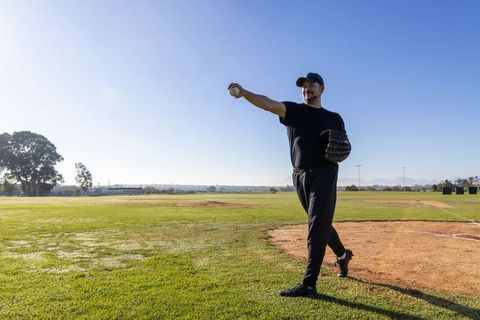 Mature man throwing baseball on sunlit pitching mound on open green field wearing glove