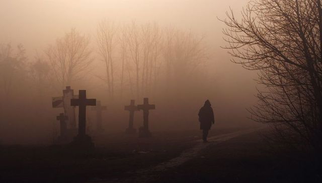 Solitary hooded figure walking through amber fog in cemetery with silhouetted crosses