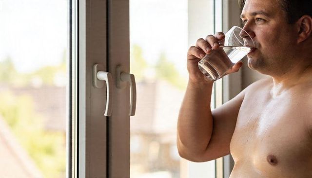 Shirtless man drinking water by bright window during home hydration and summer cool-down