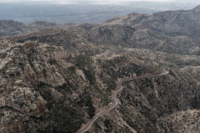 Aerial view of a winding road cutting through a rugged mountain landscape in an arid desert region. The rocky terrain is sparse with vegetation, emphasizing the arid climate. Ideal for use in travel blogs, tourism brochures, adventure magazines, and nature documentaries showcasing scenic routes and mountainous landscapes.