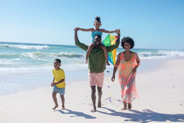 African American family enjoying a sunny day at the beach. Father carries daughter on shoulders, mother walks alongside, son runs joyfully. Perfect for articles on family vacations, summer activities, outdoor fun, and family bonding.