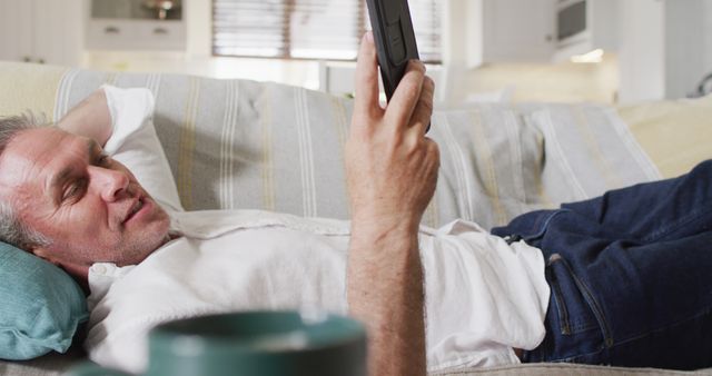 Middle-aged man relaxing on a couch at home, holding a tablet and watching a streaming video. He is dressed in casual clothes and looks comfortable and at ease. This image is perfect for showcasing technology use in home settings, leisure activities, and promoting streaming services or electronics.
