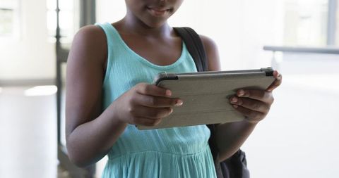 African american girl using tablet in school corridor holding folio and backpack