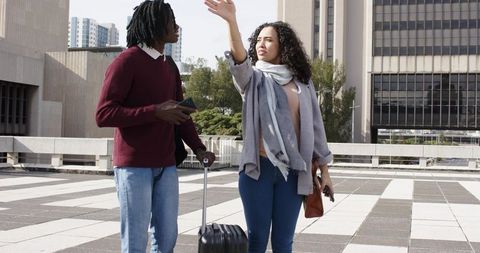 Multicultural young couple navigating city plaza with wheeled suitcase and smartphone