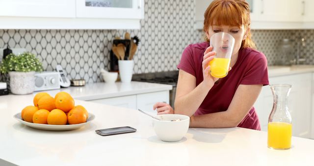 Redheaded woman drinking orange juice in a modern white kitchen with a bowl of cereal and plate of fresh oranges on the counter. Ideal for promoting healthy lifestyle, morning routines, breakfast, and modern kitchen interiors.