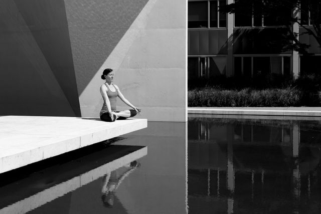 Woman sitting on a raised platform near reflective water, meditating in a black and white urban environment. She is wearing a tank top and has short hair. This can be used for promoting mindfulness, urban relaxation spaces, mental well-being, and yoga classes. Suitable for websites, posters, or articles on meditation and city living.