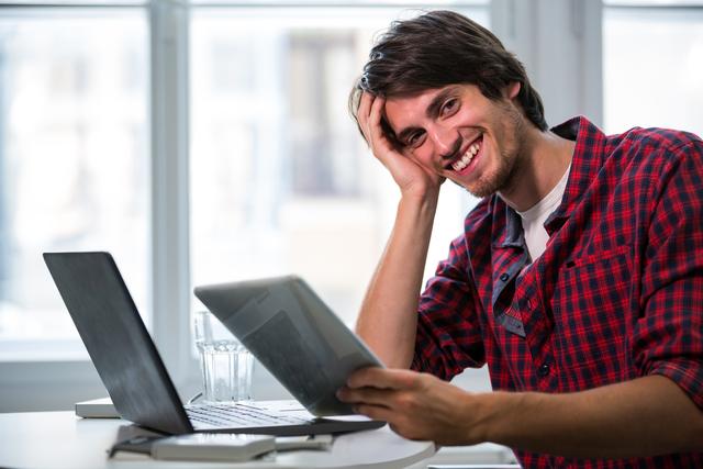 Young male executive in a casual plaid shirt smiling while holding a digital tablet in a modern office. Ideal for use in business, technology, and workplace-related content, showcasing a relaxed and confident professional environment.