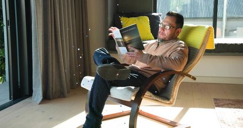 Mature man reading book relaxing in sunlit living room armchair by sliding glass doors