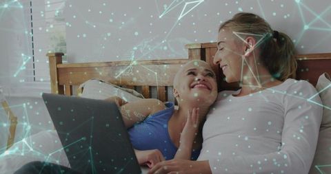Cuddling same-sex couple using laptop on bed with digital network overlay