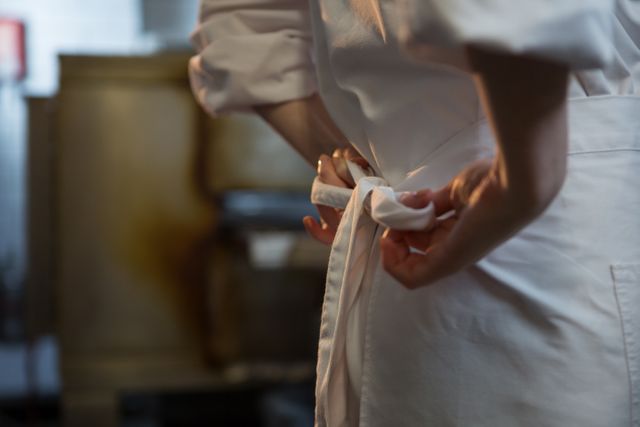 This image shows a female chef tying her apron in a professional kitchen, focusing on the mid-section. Ideal for use in culinary blogs, restaurant websites, cooking classes, and hospitality industry promotions. It emphasizes the preparation and dedication of kitchen staff.