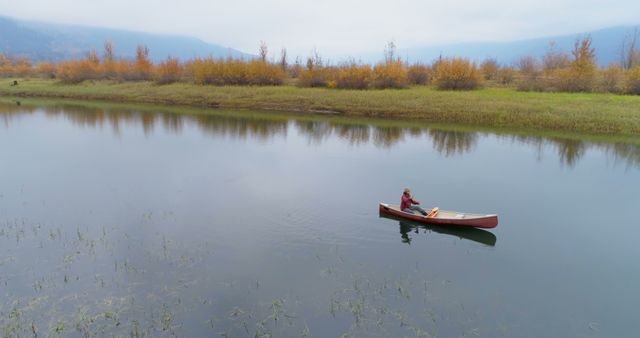 Calm lake with a single canoeist paddling leisurely amidst autumn foliage. Ideal for illustrating serenity, outdoor recreation, adventure travel, or promoting nature retreats. Can be used in advertisements for tourism, relaxation products, or environmental awareness campaigns.