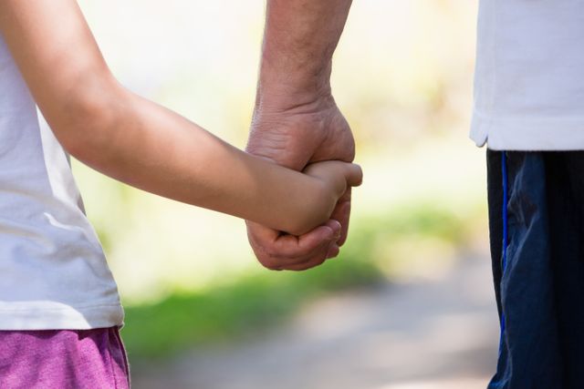 This image captures a close-up of a grandfather holding his granddaughter's hand while walking in a forest. It conveys themes of family bond, love, and support. Ideal for use in articles or advertisements about family relationships, generational connections, and outdoor activities.