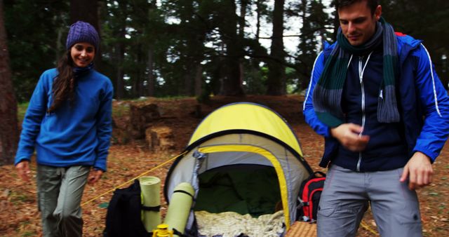 Couple dressed in warm clothing enjoying a camping trip in a forest. The scene includes a pitched tent, backpacks, and rolled-up mats. This image can be used in content related to outdoor adventures, leisure activities, camping equipment promotions, and travel blogs.
